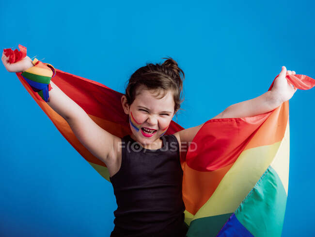 Adorable girl closing eyes while raising colorful flag above head against vibrant blue background — Stock Photo