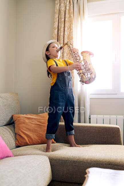 Vue latérale d'un enfant pieds nus jouant du saxophone debout sur un canapé à la maison pendant la journée — Photo de stock