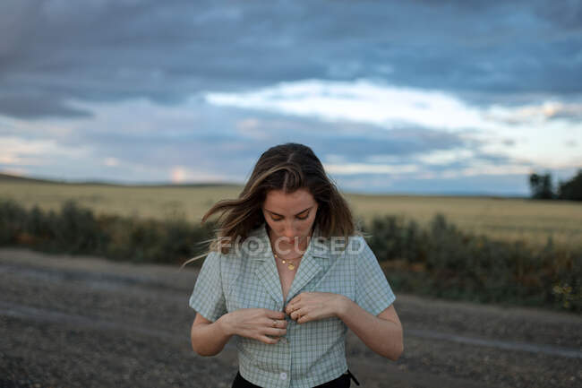Trendy young female buttoning up shirt on roadway against field under cloudy sky in twilight — Stock Photo
