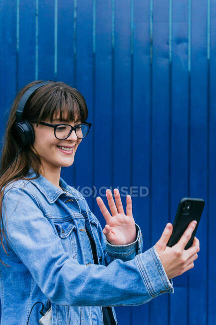 Donna positiva in cuffie con video chat su smartphone e mano ondulata mentre in piedi su sfondo blu in strada — Foto stock