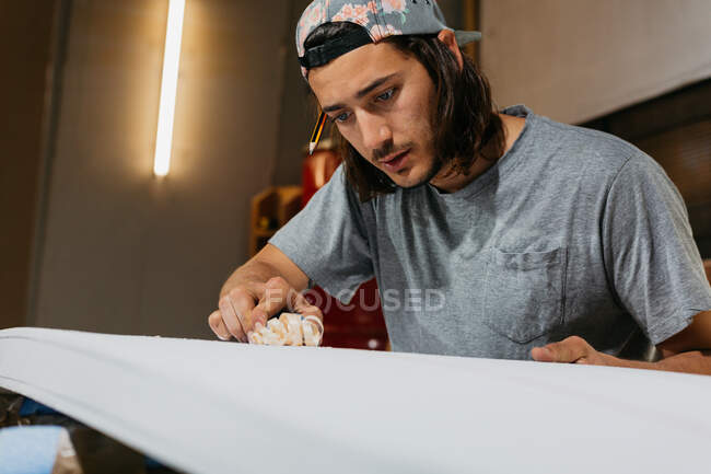 Maestro masculino enfocado usando el plano de mano y formando la superficie lisa de la tabla de surf en el taller - foto de stock