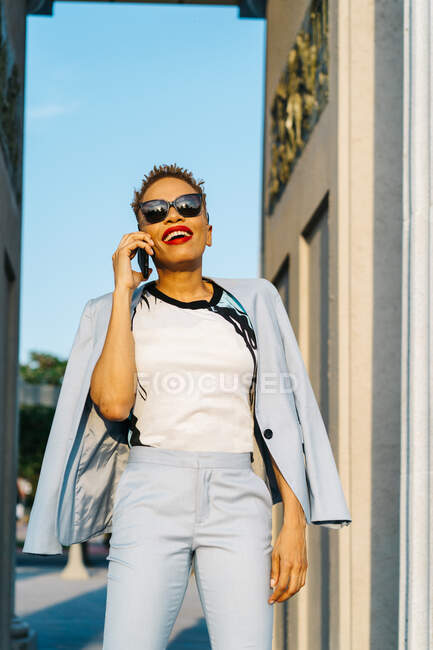 Elegante donna afroamericana adulta con taglio di capelli moderno e giacca conversare sul cellulare alla luce del sole — Foto stock