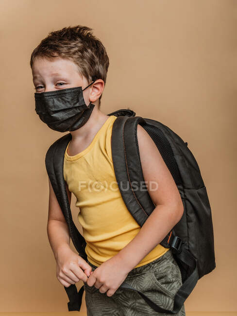 Konzentriertes Frühchen mit Rucksack und in medizinischer Schutzmaske vor Coronavirus auf braunem Hintergrund im Studio — Stockfoto