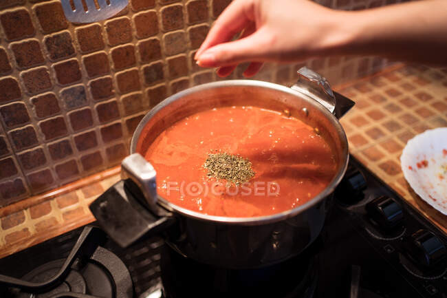 Сверху неузнаваемый урожай женский наливают ароматические специи в кастрюлю с горячим соусом маринара, сделанным из помидоров на плите в домашней кухне — стоковое фото