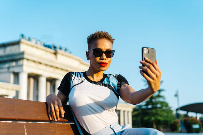 Stylish mature black female in sunglasses with red lips and modern haircut taking self portrait on cellphone in town — Stock Photo