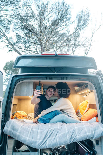 Joven pareja multirracial enamorada tomando selfie en el teléfono inteligente mientras abrazan y besan felizmente dentro de la caravana durante el viaje romántico juntos - foto de stock