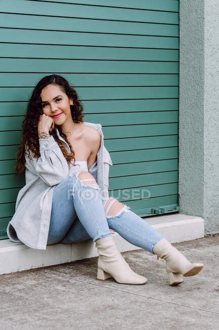 Charming female in stylish wear sitting near wall of building and leaning on hand while smiling and looking at camera — Stock Photo