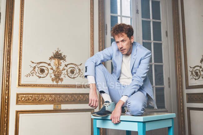 Nachdenklicher Mann im trendigen Anzug sitzt auf einem Tisch im noblen Zimmer mit Ornamenten an den Wänden, die wegschauen — Stockfoto