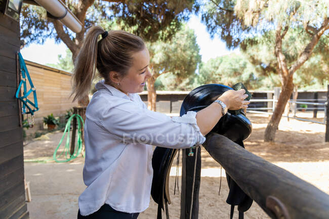Vista lateral do jóquei feminino lavar sela preta enquanto se prepara para passeios a cavalo na fazenda no verão — Fotografia de Stock