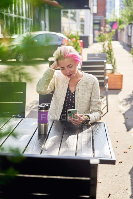 Alternatives Weibchen mit kurzen Haaren surft auf dem Smartphone in den sozialen Medien, während sie an sonnigen Tagen am Tisch im Straßencafé sitzt — Stockfoto