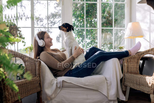 Вид сбоку веселой женщины в наушниках, развлекающейся с породистой собакой в кресле у окна в доме — стоковое фото