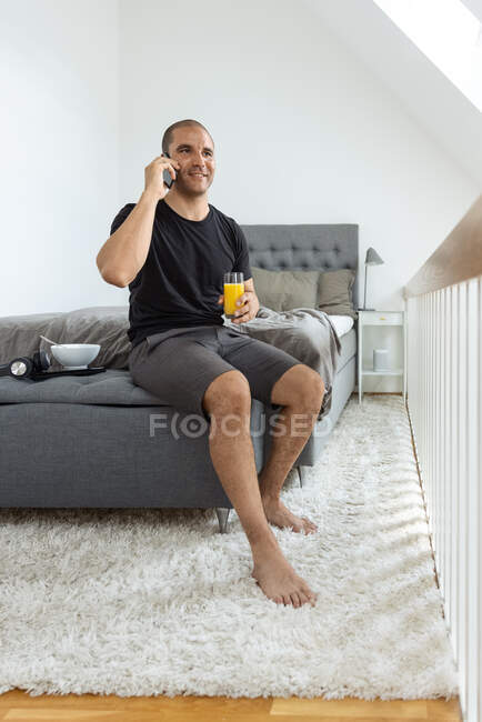 Hombre alegre sentado en la cama y hablando por teléfono móvil durante el desayuno en la mañana en casa - foto de stock