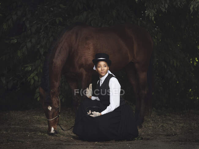 Fiduciosa signora afroamericana adulta in abiti eleganti e cappello seduto con cavallo marrone, mentre guardando lontano vicino agli alberi di giorno — Foto stock