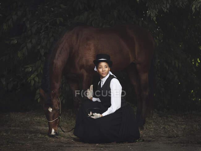 Selbstbewusste erwachsene Afroamerikanerin in eleganter Kleidung und Hut sitzt mit braunem Pferd und schaut tagsüber in der Nähe von Bäumen weg — Stockfoto