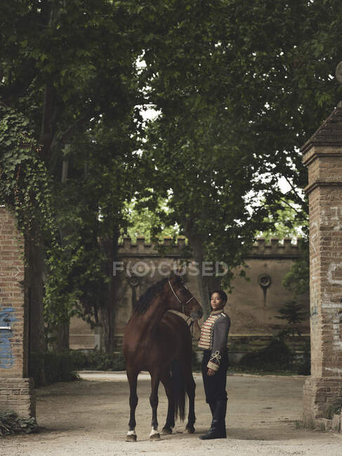 Full length of adult black female in elegant outfit standing with brown horse near green trees and castle fence in daytime in yard — Stock Photo