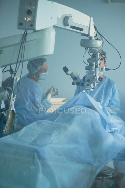 Medico anonimo in uniforme sterile che guarda attraverso il microscopio sopra il paziente sul letto contro l'assistente femminile durante l'intervento in clinica — Foto stock
