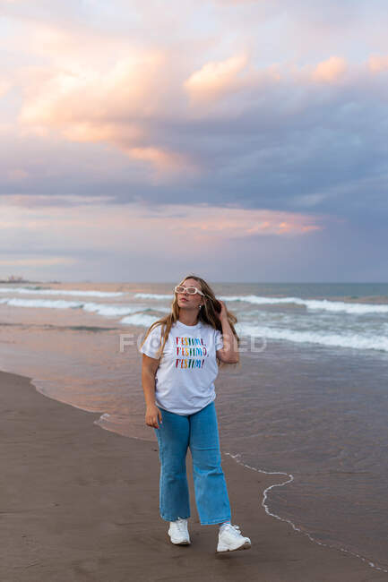 Позитивная молодая женщина в модных солнцезащитных очках и стильной одежде, прогуливающаяся по морю в летний вечер — стоковое фото