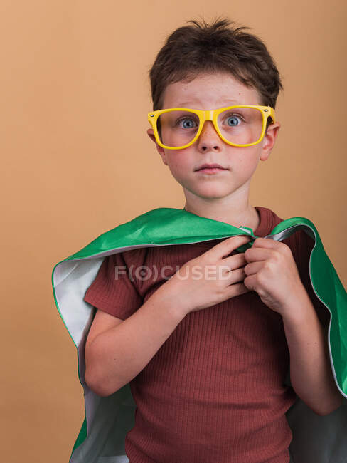 Schockiertes Kind mit blauen Augen in Plastikbrille und Heldenumhang blickt in die Kamera auf beigem Hintergrund — Stockfoto