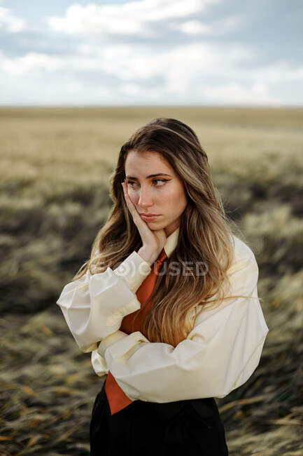 Безразличная молодая женщина в белой рубашке с красным галстуком касается щеки, глядя на сельхозугодия — стоковое фото