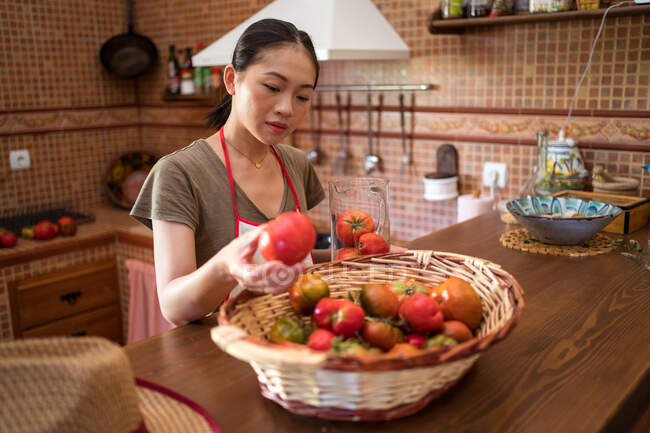 Femme au foyer ethnique ciblée sélectionnant des tomates fraîches sur la cuisine tout en cuisinant des aliments à la maison — Photo de stock