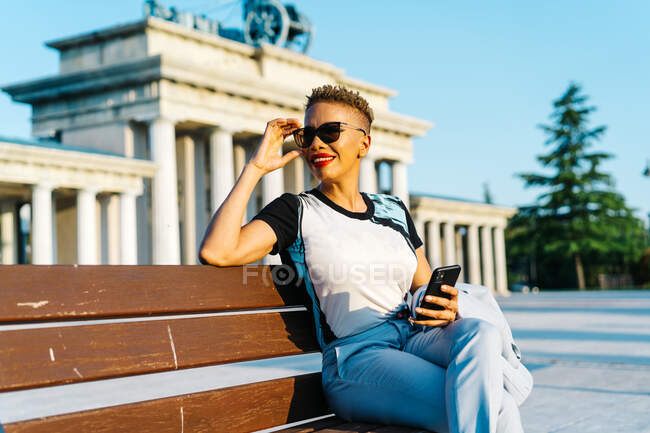 Mujer étnica de moda en gafas de sol con teléfono inteligente y corte de pelo moderno mirando hacia otro lado mientras se sienta en el banco de la ciudad - foto de stock