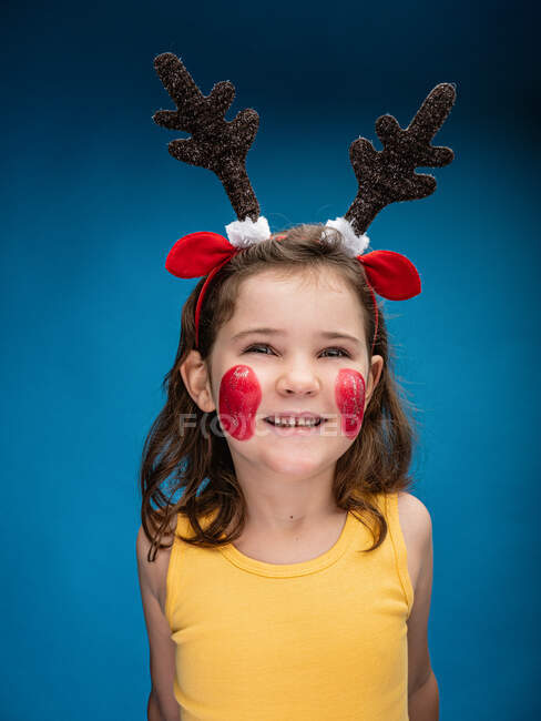 Улыбающаяся девушка с покрашенными в красный цвет щеками, в игрушечные оленьи рога и уши и смотрит в камеру на синем фоне — стоковое фото
