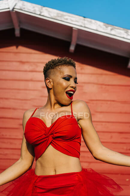Femme afro-américaine à la mode avec lèvres rouges et cheveux courts dansant tout en criant contre le mur de la maison — Photo de stock