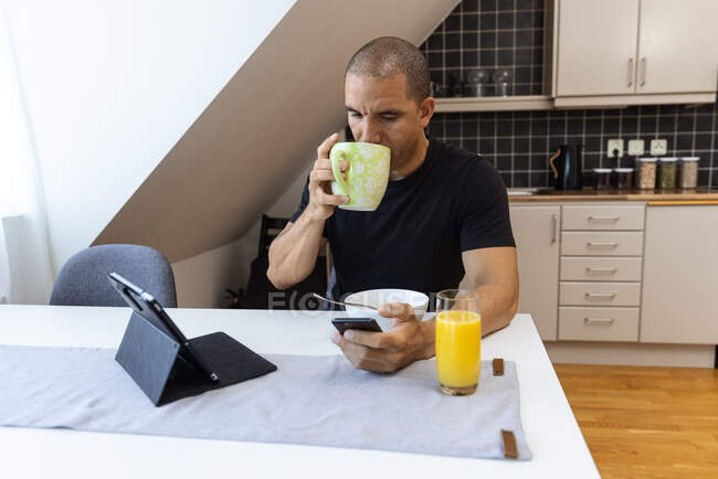 Дорослий чоловік п'є гарячий напій з чашки та переглядає мобільний телефон, сидячи за столом на кухні та снідаючи вранці вдома — стокове фото