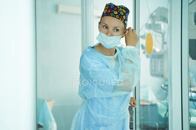 Adult female doctor in surgical uniform and ornamental medical cap putting on disposable mask while looking forward in hospital — Stock Photo