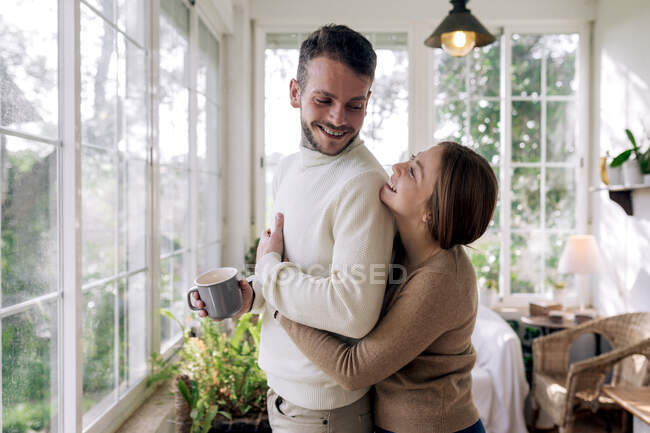 Вид збоку весела жінка, яка обіймає бородатого чоловіка, улюбленого чашкою кави, дивлячись у вікно в будинку — стокове фото
