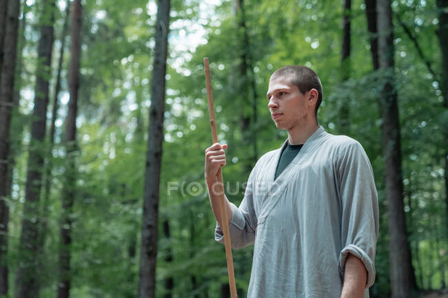 Uomo con bastone che tiene la mano vicino al petto mentre pratica il kung fu nel bosco — Foto stock