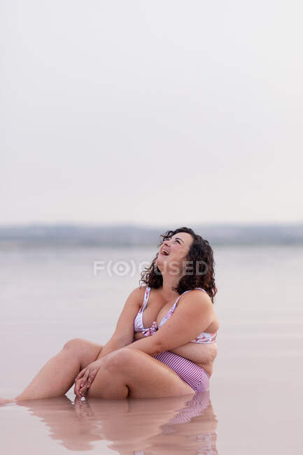 Entzückte kurvige Frau im Bikini sitzt im Wasser des rosa Teiches im Sommer und schaut auf — Stockfoto
