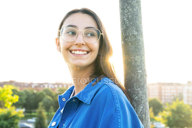 Seitenansicht einer fröhlichen Frau in stylischem Outfit, die sich auf einen Baumstamm lehnt, während sie im Gegenlicht auf dem grünen Rasen gegen die Stadt steht — Stockfoto
