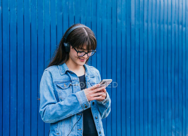 Mulher positiva em fones de ouvido navegando no smartphone enquanto estava em fundo azul na rua — Fotografia de Stock