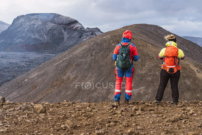 Senderistas de pie en la montaña contra Fagradalsfjall con lava y humo bajo el cielo nublado en Islandia - foto de stock