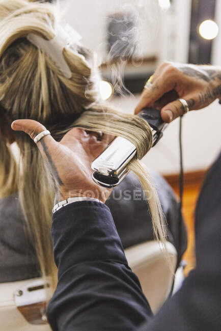 Peluquero masculino anónimo usando plancha para rizar mechones rubios de cliente femenino durante el trabajo en salón de belleza - foto de stock