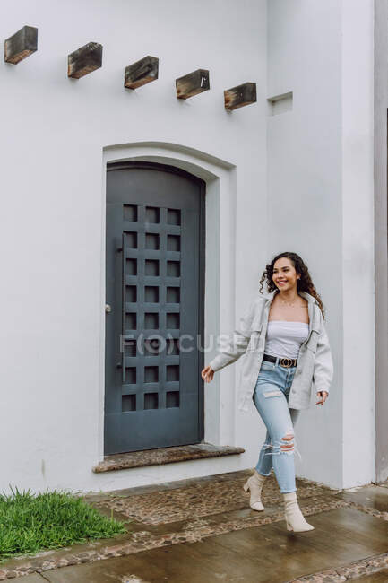 Mulher encantada em roupas da moda em pé perto da porta do edifício residencial e desfrutando de tempo chuvoso na cidade — Fotografia de Stock