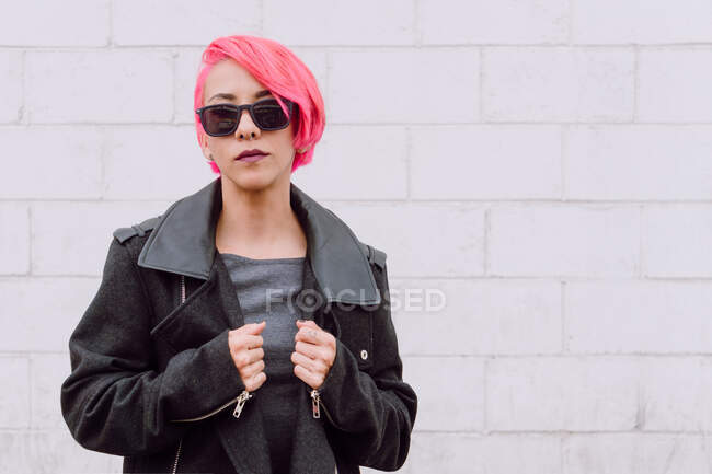 Стильная модель с розовыми волосами в модном пальто и солнцезащитных очках, смотрящая на камеру на белой кирпичной стене — стоковое фото