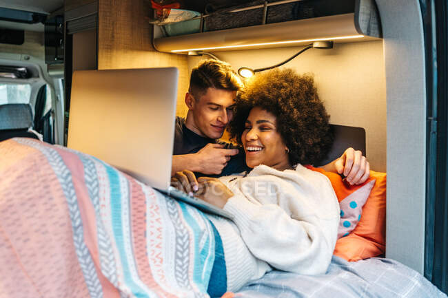 Feliz casal multirracial romântico abraçando e aproveitando o tempo juntos enquanto deitado na cama e assistindo filme no laptop durante a viagem em van campista — Fotografia de Stock