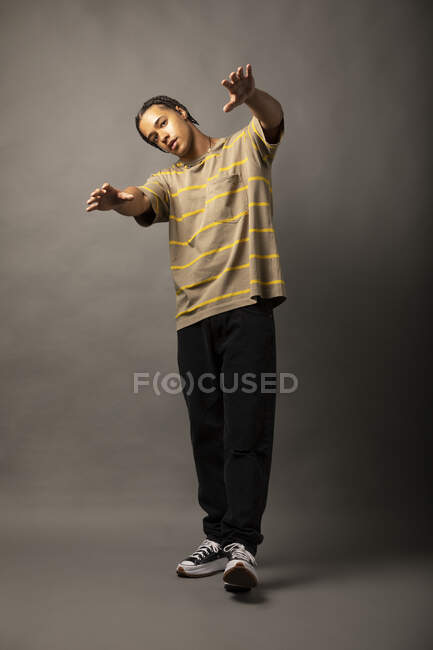 Full body of African American hipster male model with braided hair dressed in casual striped shirt and black trousers with sneakers doing dance moves against gray wall — Stock Photo
