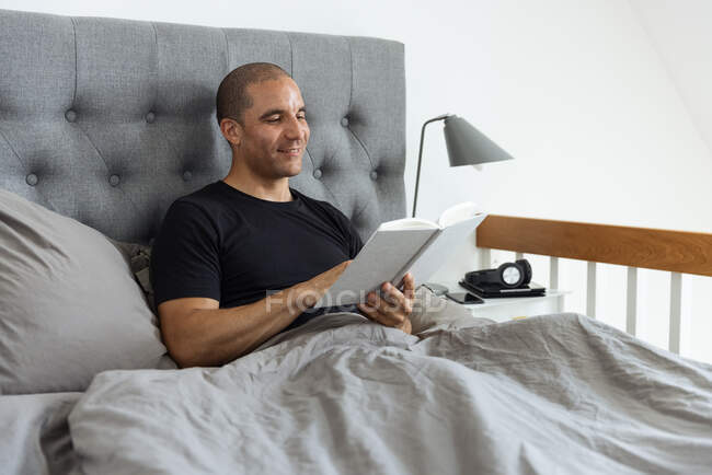 Hombre pensativo sentado en la cama blanda por la mañana y leyendo interesante historia en el libro después de despertar - foto de stock