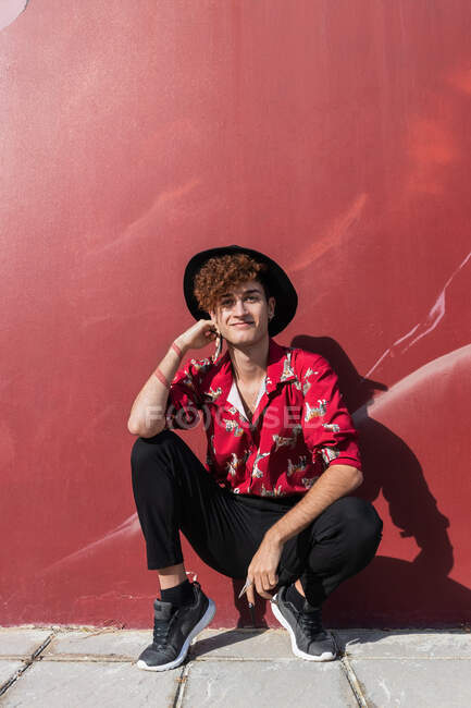 Homme homosexuel heureux à la mode avec de longs ongles en chemise ornementale regardant la caméra tout en squattant sur le trottoir contre le mur — Photo de stock