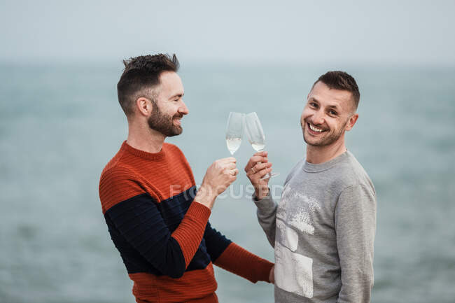 Vista lateral de alegres hombres homosexuales tintineando vasos con champán salpicado mientras mira a la cámara contra el océano - foto de stock