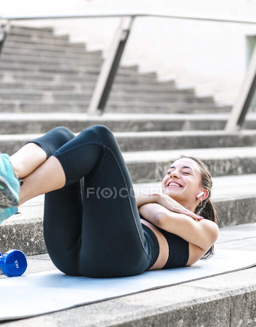 Atleta femenina feliz con las manos cruzadas en el pecho sonriendo y haciendo ejercicios abdominales en los pasos durante el entrenamiento de fitness en la calle - foto de stock