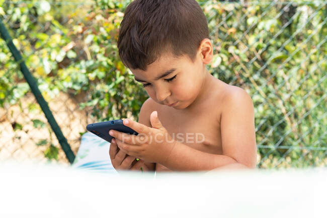 Recorte lindo niño usando el teléfono inteligente mientras está sentado en el jardín cerca de la cerca verde en el día de verano soleado - foto de stock