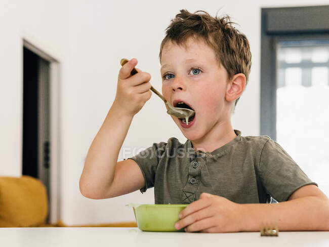 Menino adorável comendo sopa de creme apetitosa com colher e boca aberta durante o almoço em casa — Fotografia de Stock