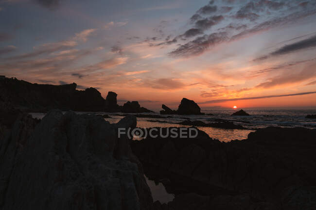 Increíble paisaje tranquilo de puesta de sol sobre el mar ondulado ondulado con rocas bajo el cielo nublado colorido en la noche de verano en Liencres Cantabria España - foto de stock
