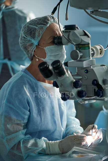 Medico donna adulto concentrato in maschera sterile e cappuccio medico ornamentale guardando attraverso il microscopio chirurgico contro il collega di coltura in ospedale — Foto stock