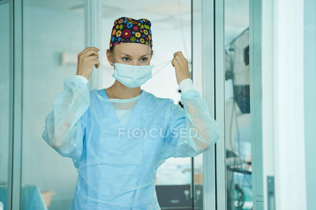Doctora adulta en uniforme quirúrgico y gorra médica ornamental que se pone una máscara desechable mientras espera en el hospital - foto de stock