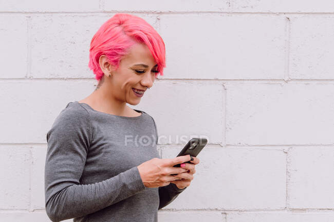 Young female with bright pink hair in casual clothes smiling and using smartphone while standing near white wall on street — Stock Photo